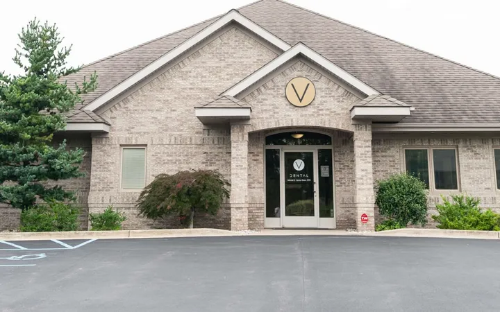 Exterior of a single-story brick dental practice with a round V sign above the entrance, landscaping and a parking lot in front