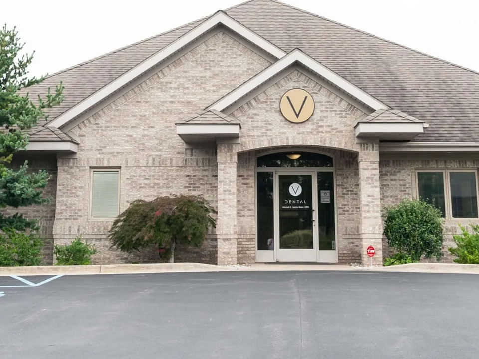 Exterior of a single-story brick dental practice with a round V sign above the entrance, landscaping and a parking lot in front