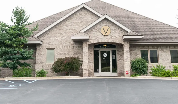 Exterior of a single-story brick dental practice with a round V sign above the entrance, landscaping and a parking lot in front