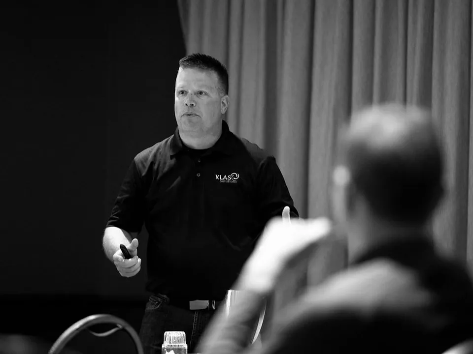 Phil Cole in a KLAS Solutions polo presenting to a seated audience, in black and white