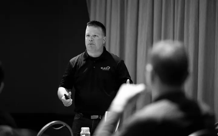 Phil Cole in a KLAS Solutions polo presenting to a seated audience, in black and white