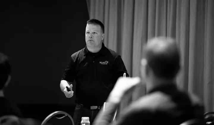 Phil Cole in a KLAS Solutions polo presenting to a seated audience, in black and white